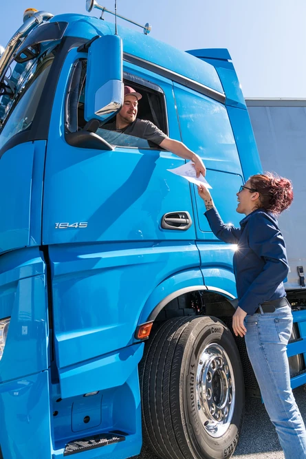 Ein LKW-Fahrer reicht einer Frau, die neben dem LKW steht, durch das Fenster ein Blatt Papier